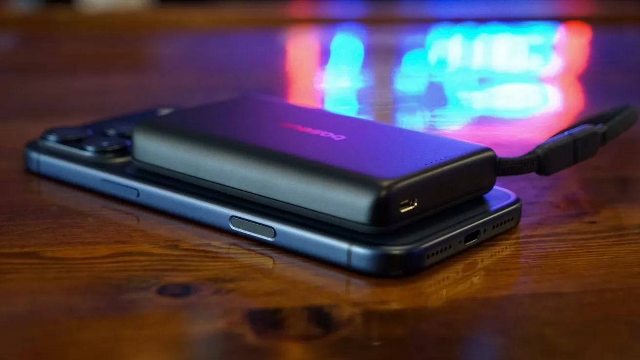 Black power bank resting on an iPhone 17 Pro Max on a wooden table, with a short cable attached and colorful blurred neon lights glowing in the background.
