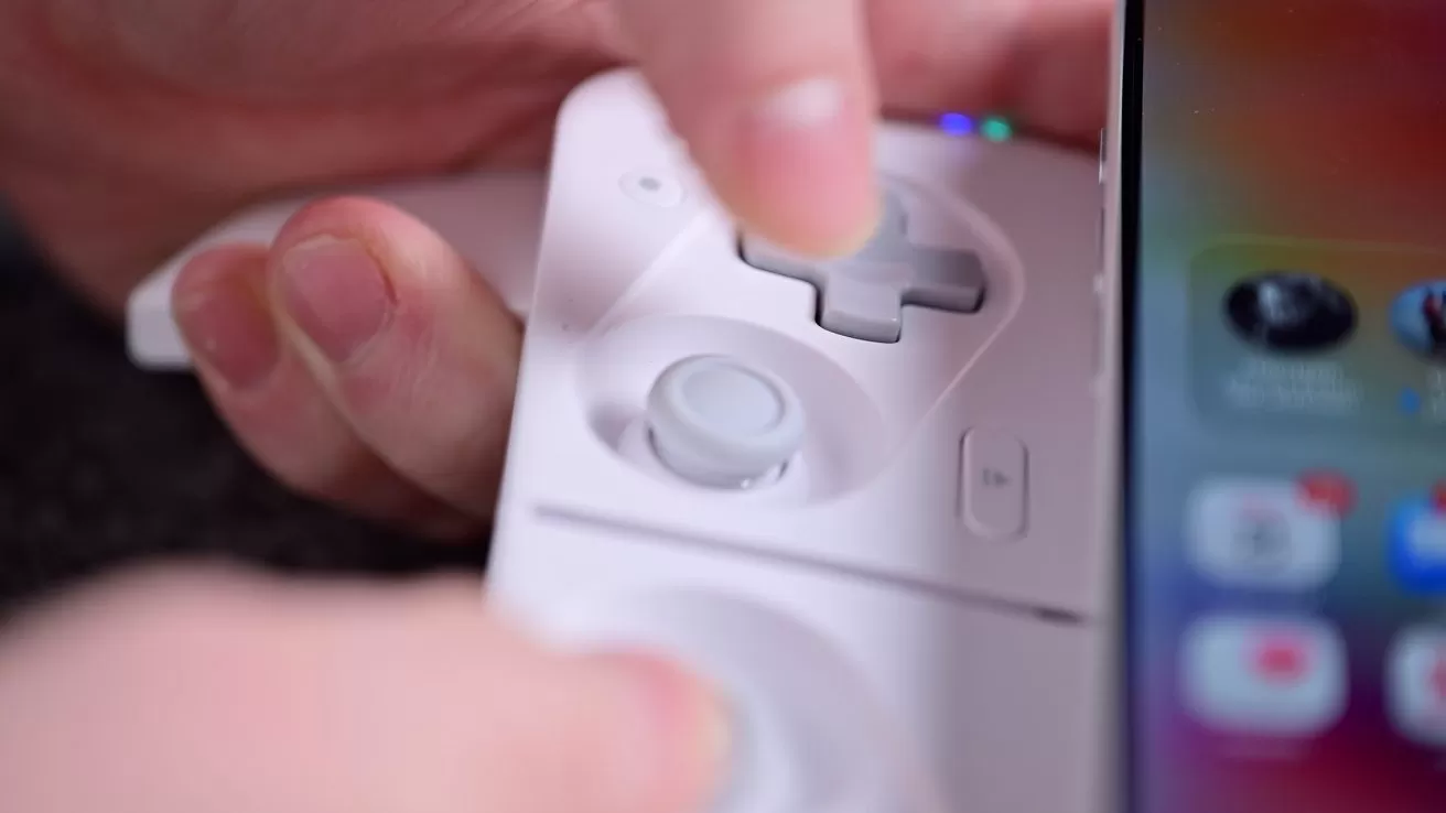 Hand holding a white gaming controller with a thumb on the directional pad, next to a phone screen displaying app icons.