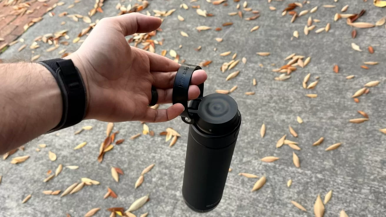 A hand with a black wristband holds a black water bottle by its loop on a concrete surface with scattered leaves.