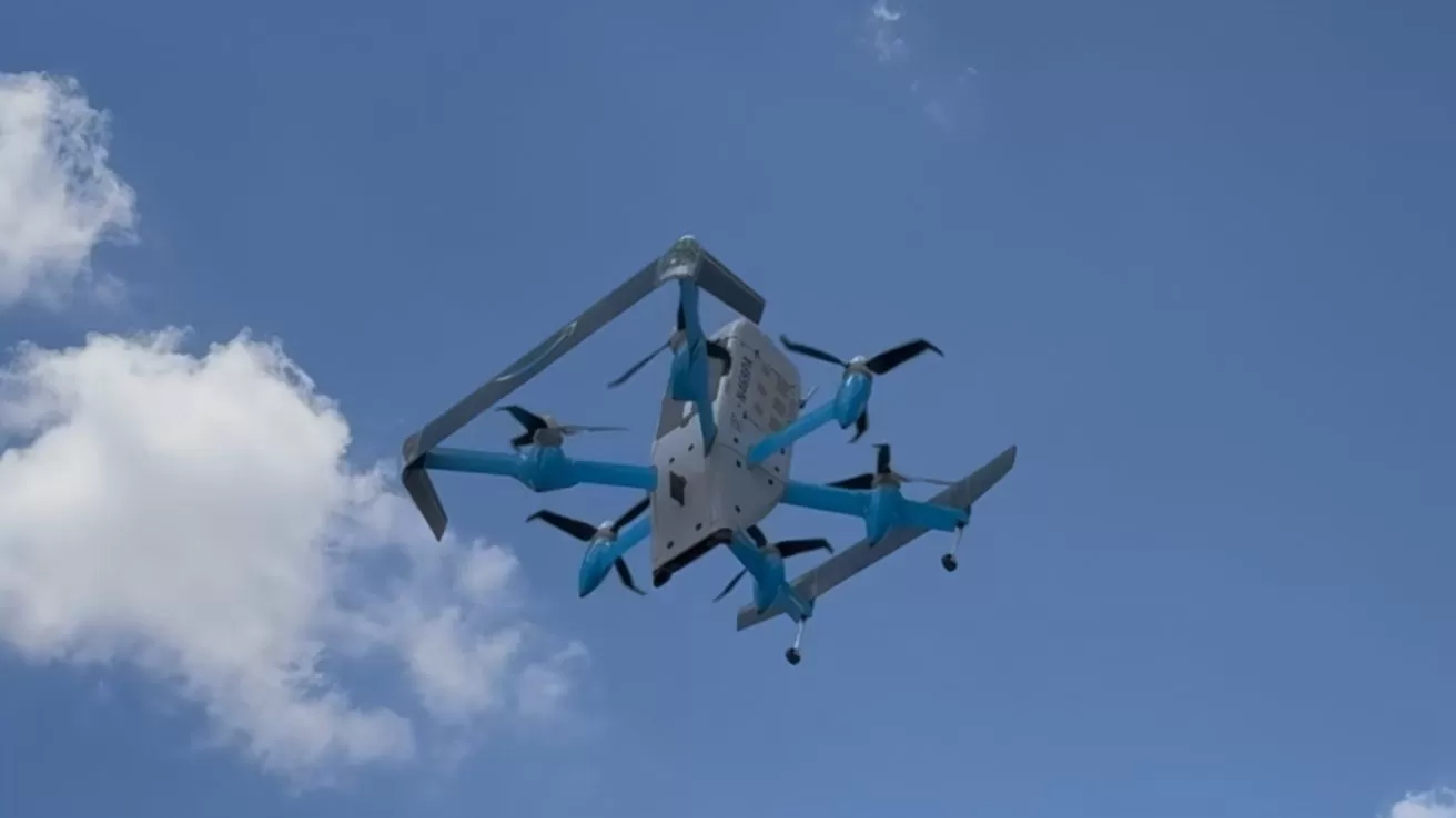 Large blue-and-white cargo drone with multiple rotors flying in a clear blue sky, viewed from below, with a few fluffy white clouds in the background