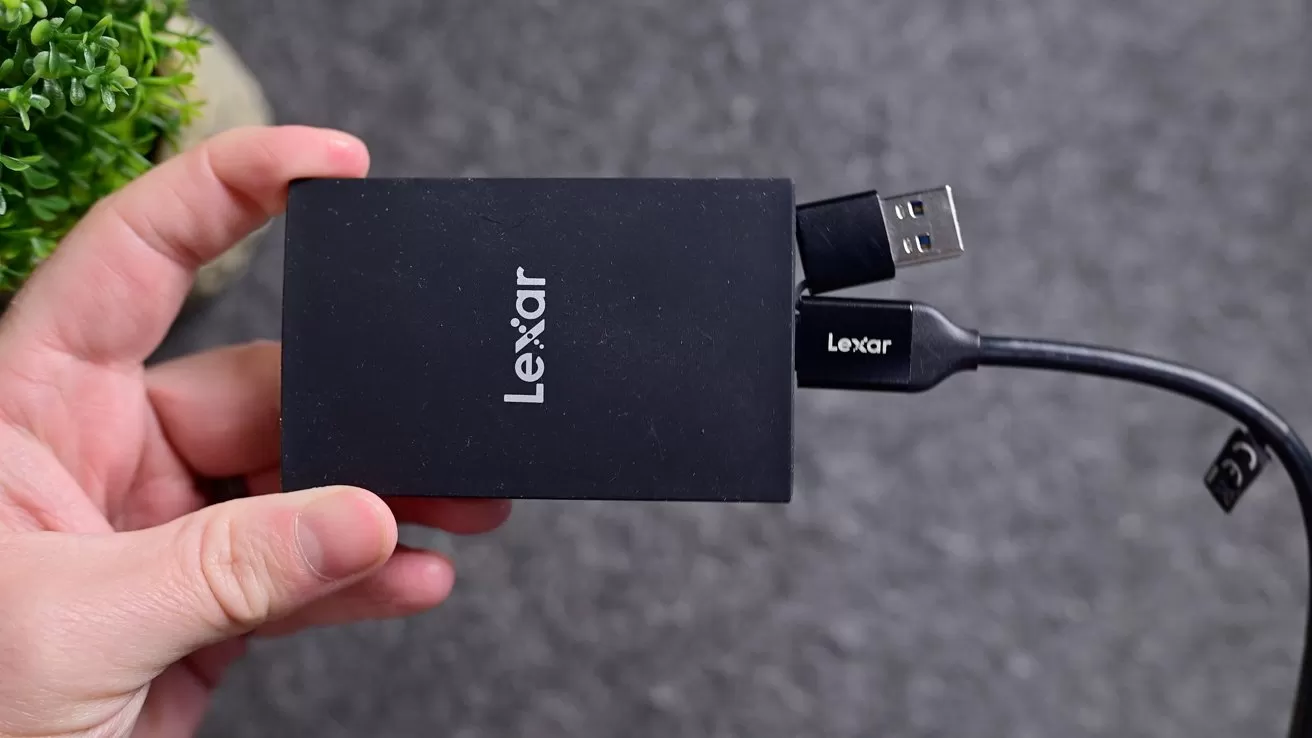 Hand holding a small black Lexar external drive with an attached USB cable, against a gray background with a green potted plant partially visible on the left