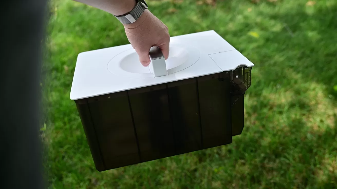 Hand holding a small rectangular box with a white lid and handle, carried outdoors above green grass, suggesting portable equipment or container being moved across a lawn