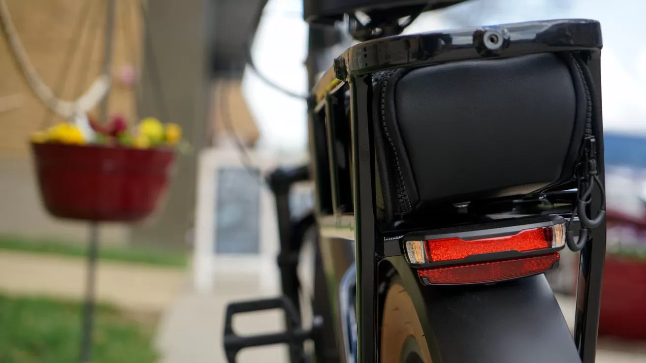 Close-up of a black bicycle rear rack with padded bag and red reflector, with blurred background showing a hanging flower pot and outdoor walkway