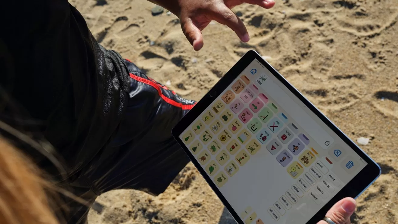 Person standing on sandy beach holding a tablet displaying colorful communication symbols and icons, viewed from above with one hand poised above the screen