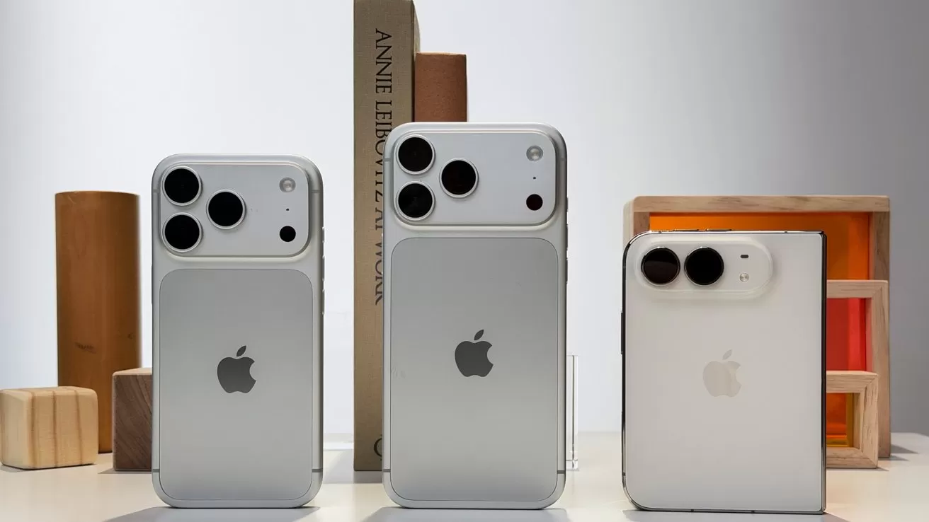 Three silver iPhones with large dual rear cameras stand upright on a table, surrounded by minimalist wooden blocks and a vertical book in soft, neutral lighting.