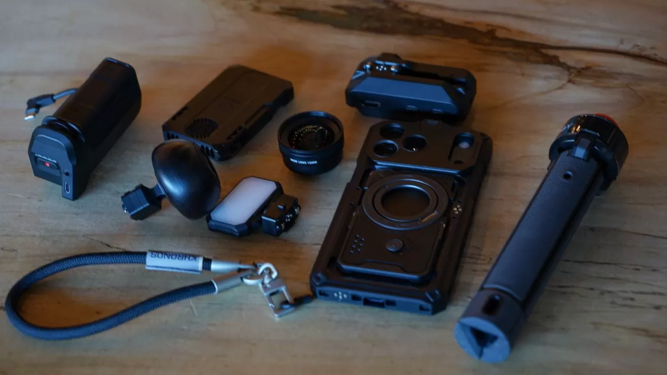 Collection of black camera accessories on a wooden surface, including rugged phone case, detachable lenses, compact lights, cylindrical grip or handle, small gadgets, and a wrist strap labeled Sandmarc