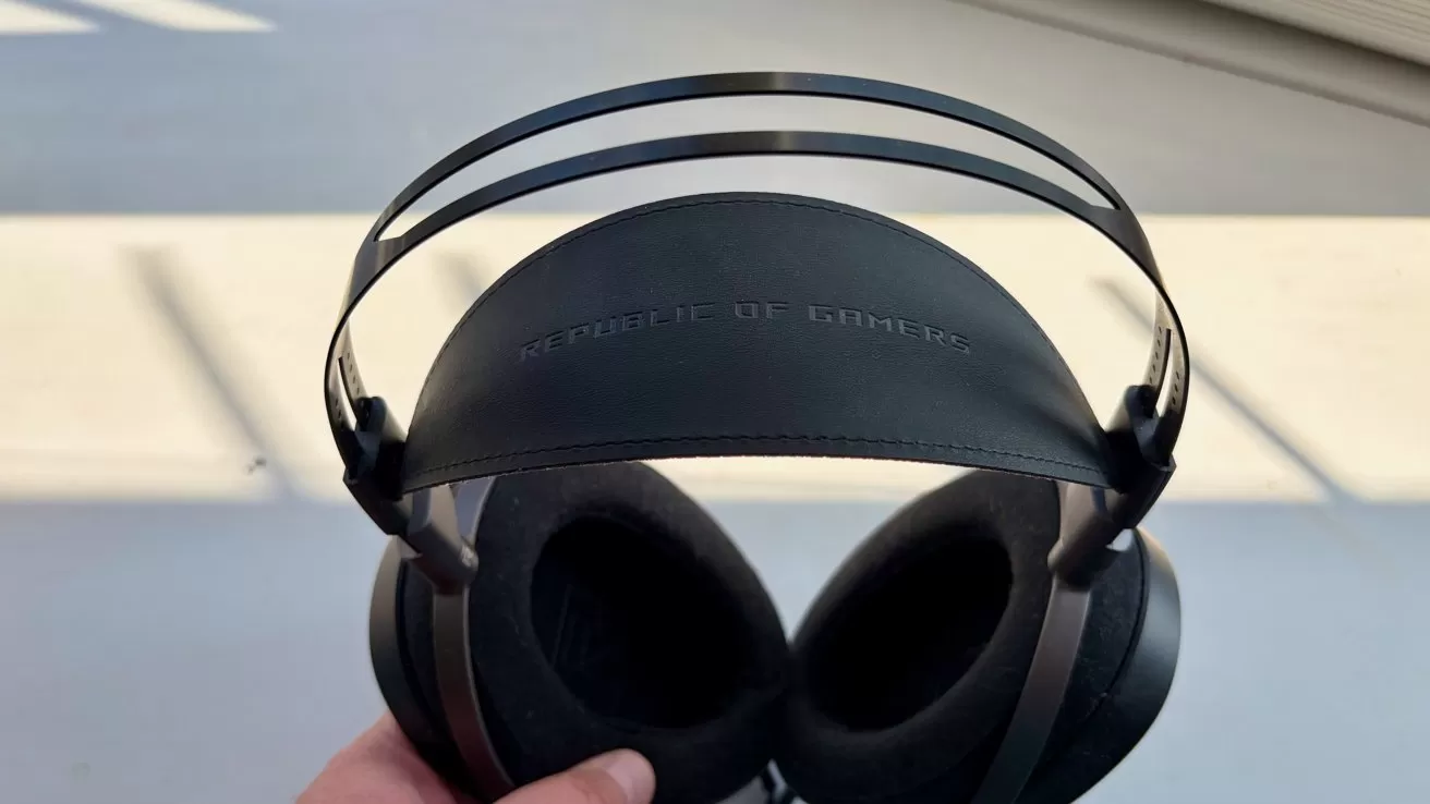 Hand holding a pair of black over-ear headphones with a wide padded headband and dual metal support bands, shown against a bright indoor background with strong sunlight and shadows