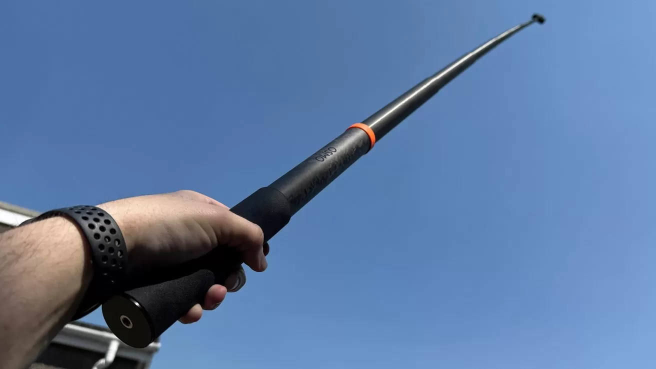 Person holding a long black telescopic pole with an orange ring, extending upward toward a clear blue sky, viewed from below with a wristband visible on their arm