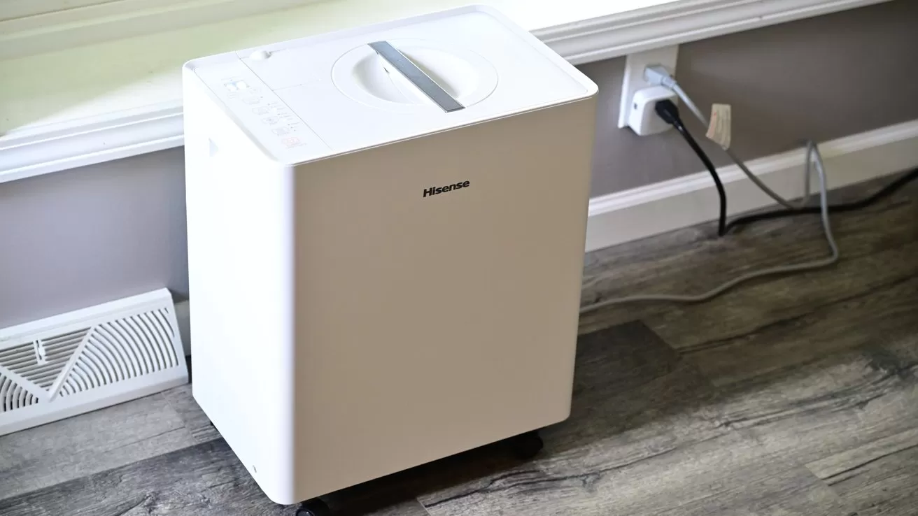 White portable dehumidifier on wheels sitting on a wood floor near a wall, plugged into an electrical outlet beside a floor vent and baseboard trim.