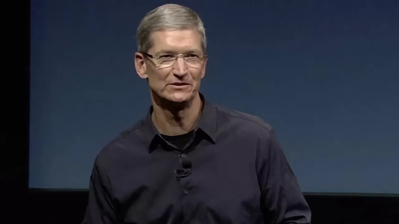 Middleaged man with short gray hair and glasses speaks onstage, wearing a dark buttondown shirt, against a plain dark blue background.