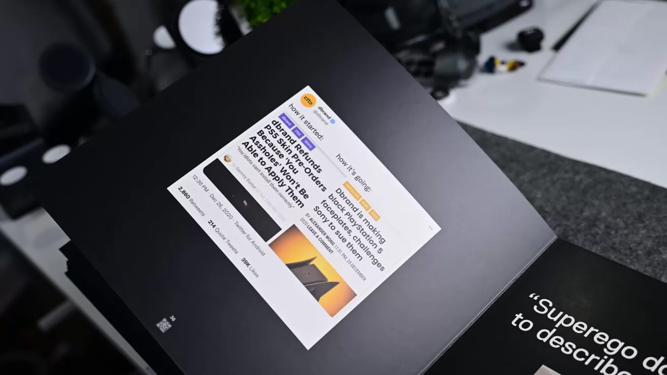 Open book on a desk displaying a printed social media post with text and a small photo, surrounded by office items like a keyboard, mouse, and camera equipment