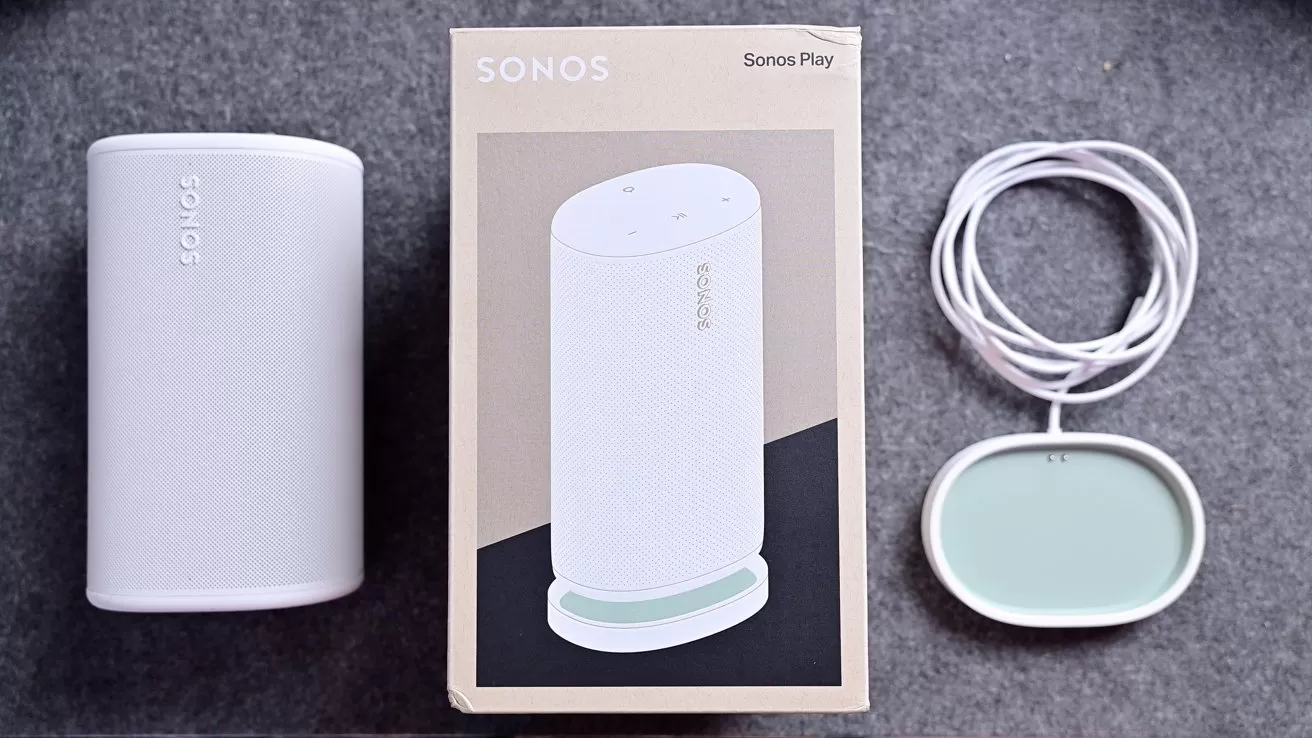 Sonos smart speaker set on gray surface, showing the cylindrical speaker, its retail box with product photo, and a small oval accessory with attached white cable