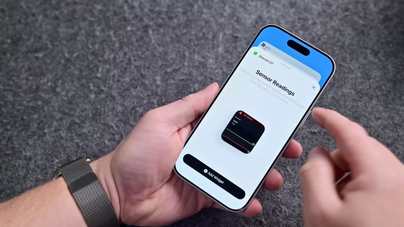 Person holding a blue smartphone above gray fabric, setting up a sensor reading widget on the screen, with another hand poised to tap, wearing a dark watchband on the wrist