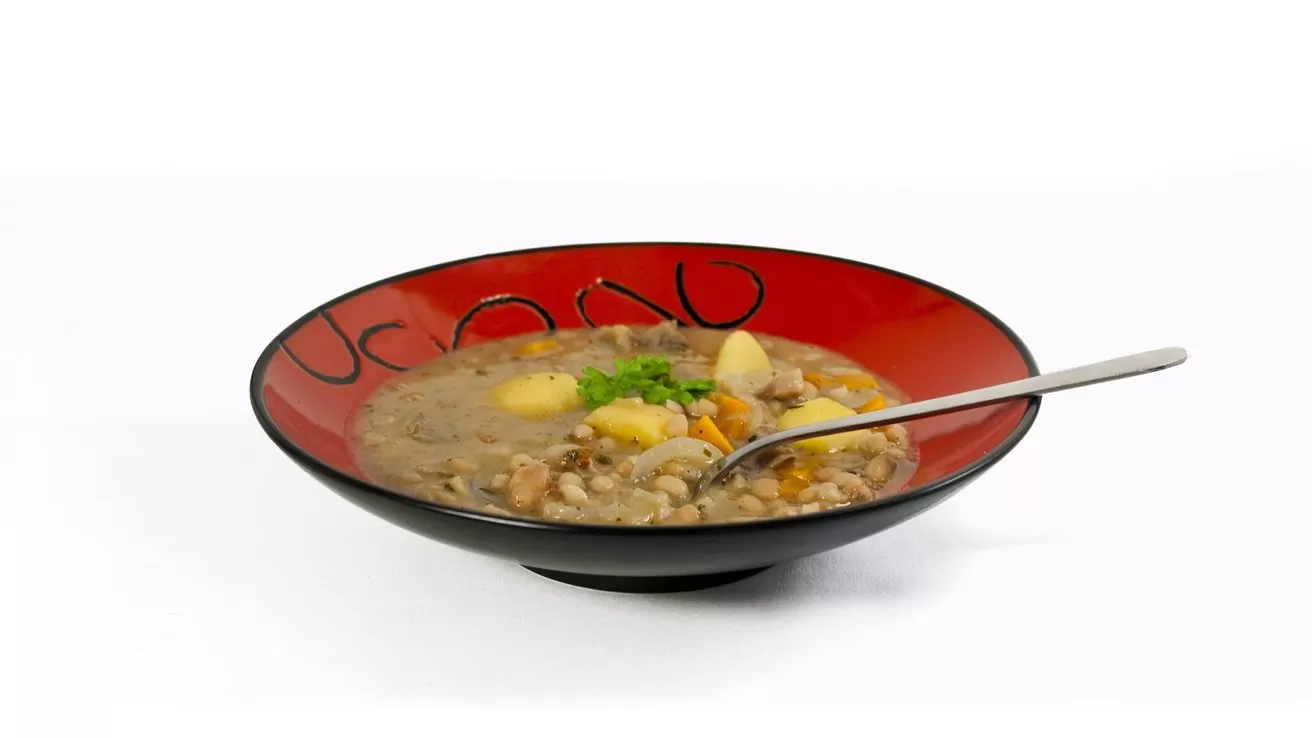 Red and black bowl of hearty lentil and vegetable stew with potatoes and carrots, topped with green herbs, with a metal spoon resting inside on a white background
