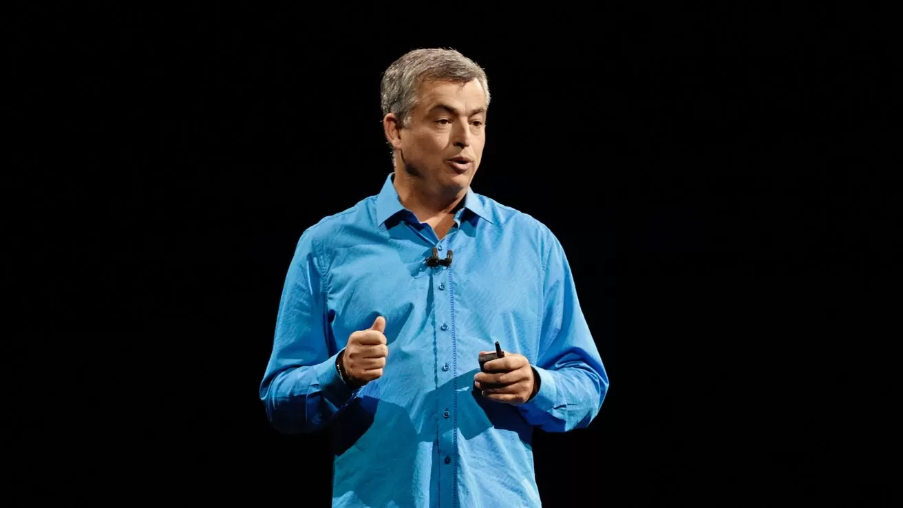 Middleaged man in a bright blue shirt speaking on stage, holding a small device, with a microphone clipped to his collar against a dark background.