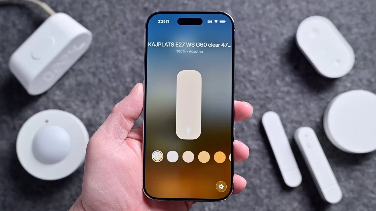 Hand holding a smartphone showing a light control app with brightness slider and color temperature options, surrounded by several small white smart home devices on a gray surface