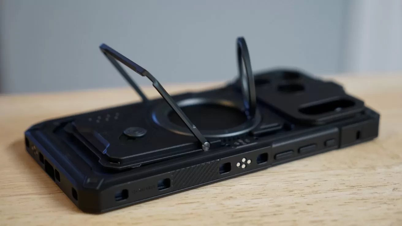 Black rugged iPhone case lying on a wooden surface, featuring a fold-out metal kickstand and circular ring stand, with cutouts for buttons, ports, and rear camera module