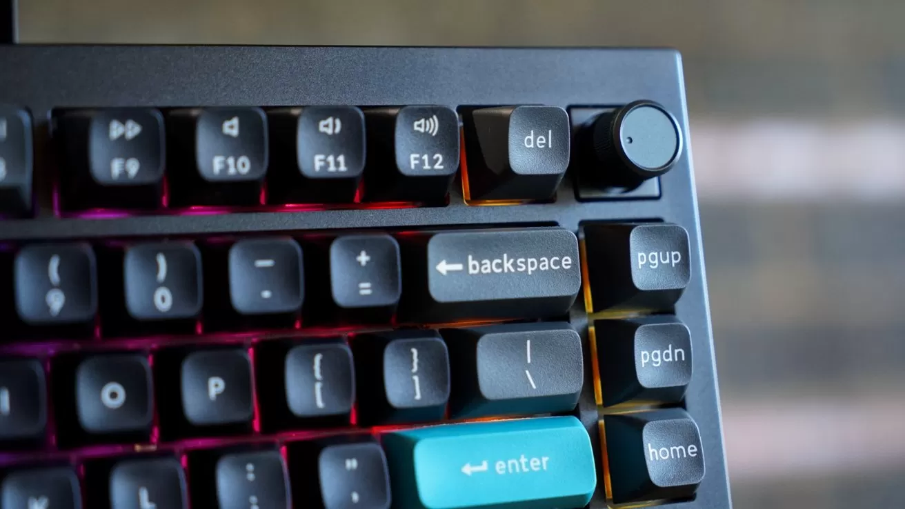 Close-up of a mechanical keyboard corner with RGB backlighting, showing backspace, teal enter, navigation keys, a delete key, and a black volume control knob on the top right