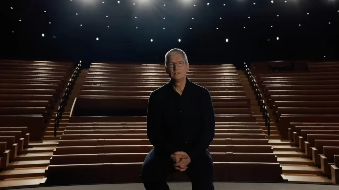 Man in dark clothing sits alone on a theater stage, hands clasped, facing forward, with empty tiered seats behind him and soft overhead spotlights glowing in the darkness