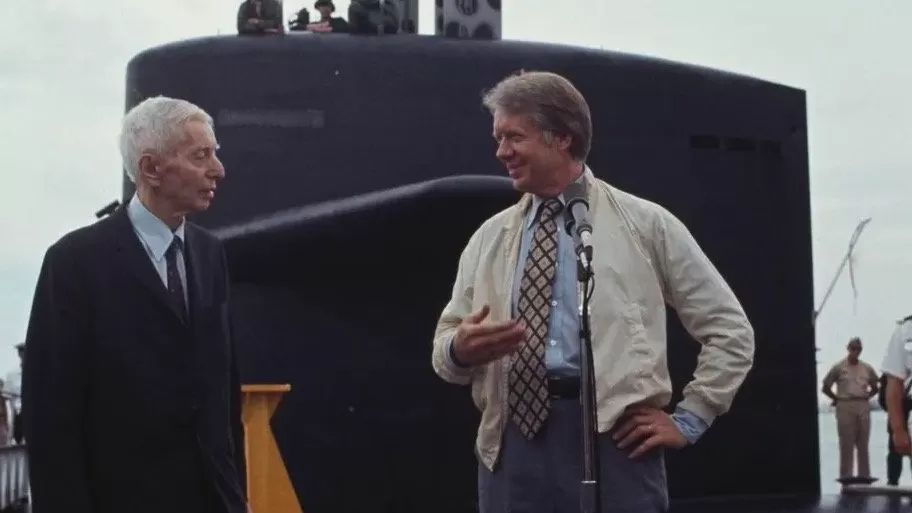 Two men stand talking in front of a large dark submarine; one wears a suit, the other a casual jacket with tie, speaking at a microphone while sailors watch in the background