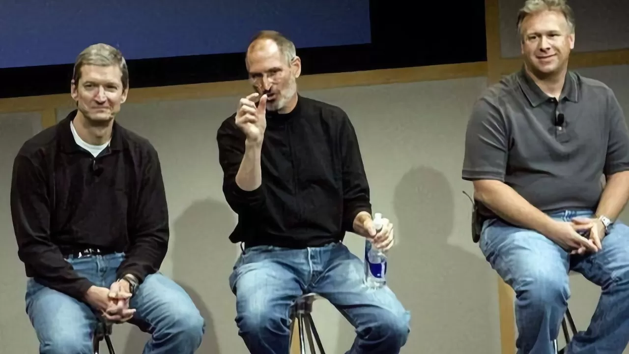 Three men sit on stools on a stage; the middle man gestures while holding a water bottle, and all wear casual clothes with jeans and microphones attached
