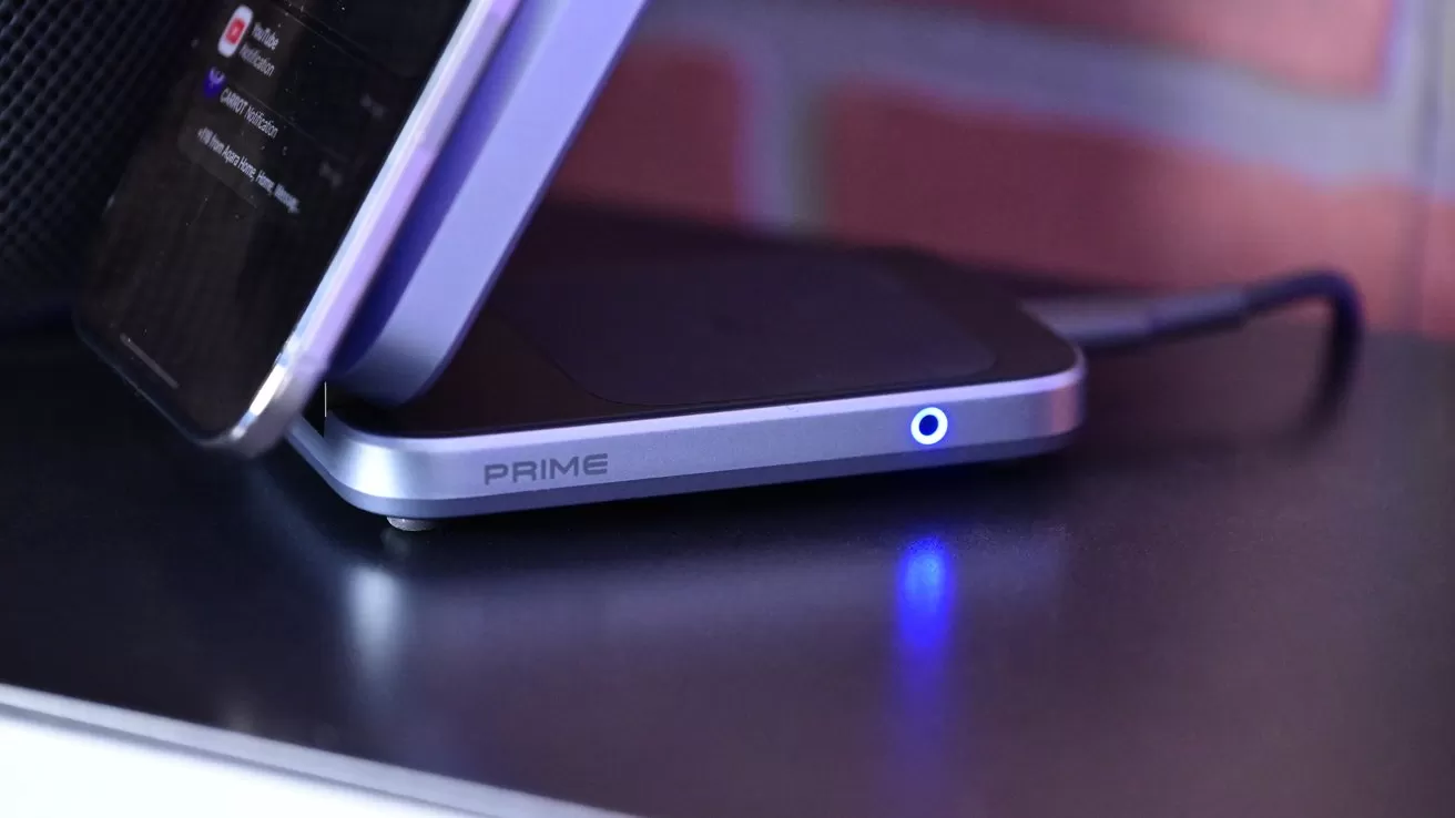 Smartphone leaning on a sleek PRIME-branded wireless charging stand, connected by a cable, with a glowing blue indicator light, set against a blurred brick wall background