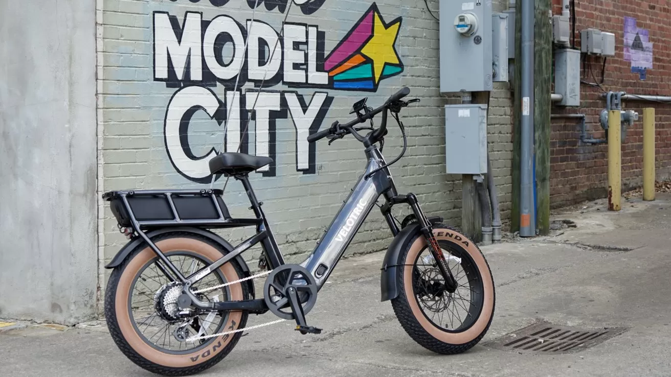 Black Velotric electric bike with fat tan tires parked in an alley beside a brick wall mural reading Model City with colorful star graphic and nearby utility boxes