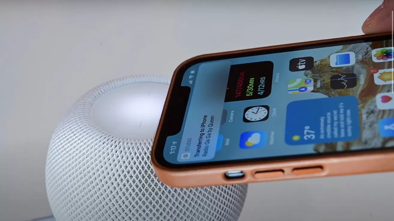 Smartphone held close to a small round smart speaker, showing the phone's home screen and suggesting a connection or interaction between the two devices