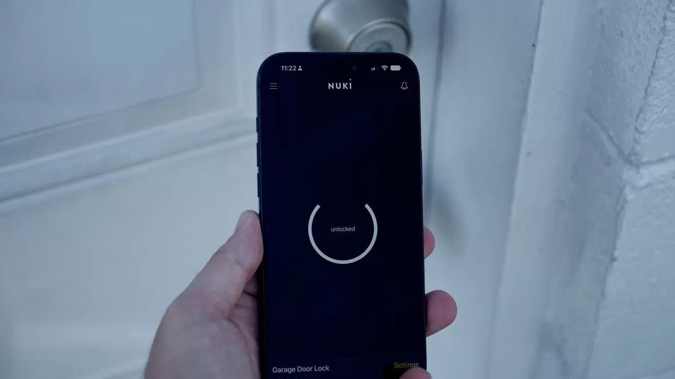 Hand holding a smartphone with a smart lock app open in front of a closed white door and round metal doorknob, suggesting remote control of the door lock