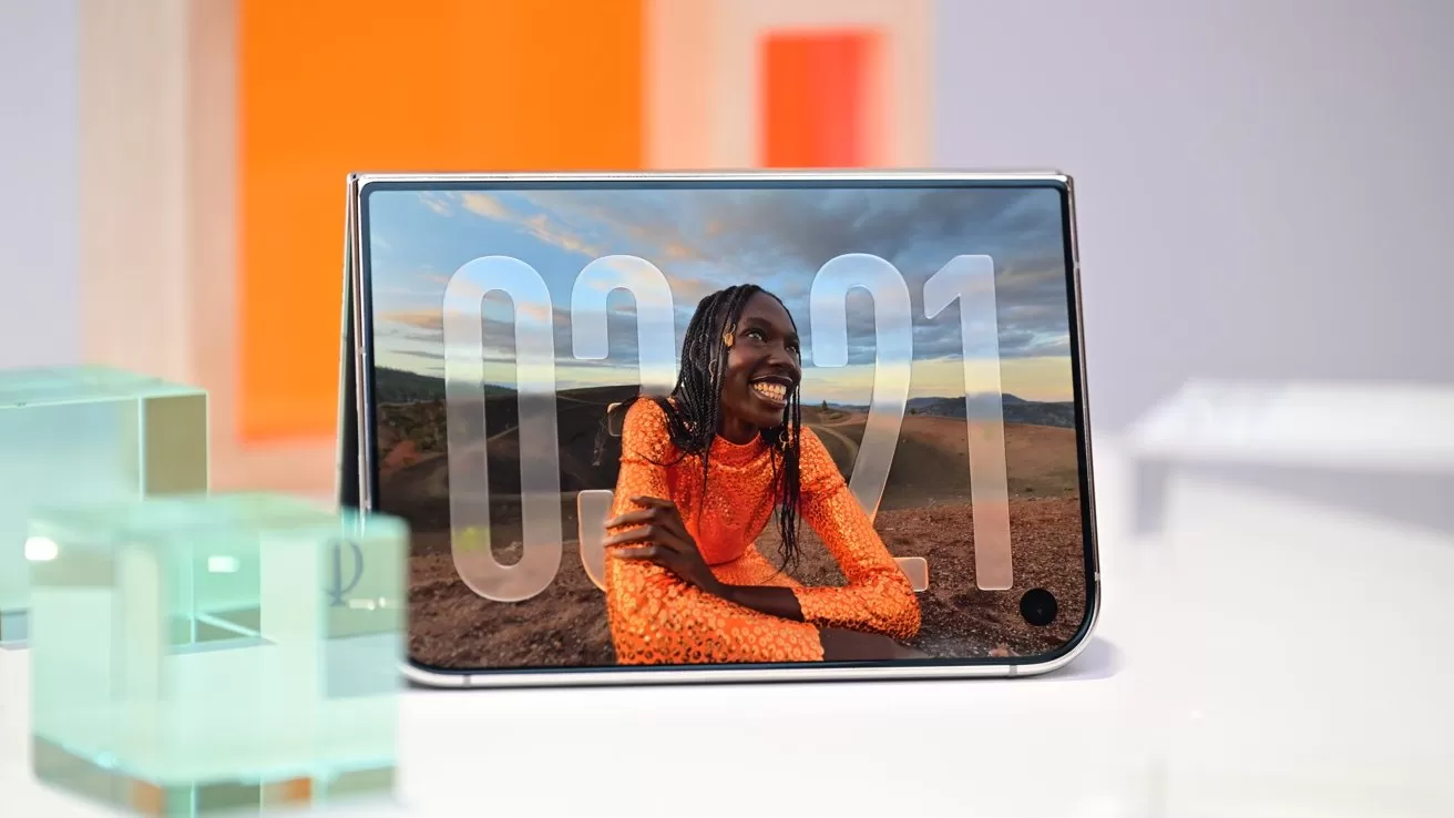 Folded smartphone on a table displaying a smiling person in an orange sweater outdoors at sunset, with large translucent numbers overlaid on the screen and colorful geometric decor in the background