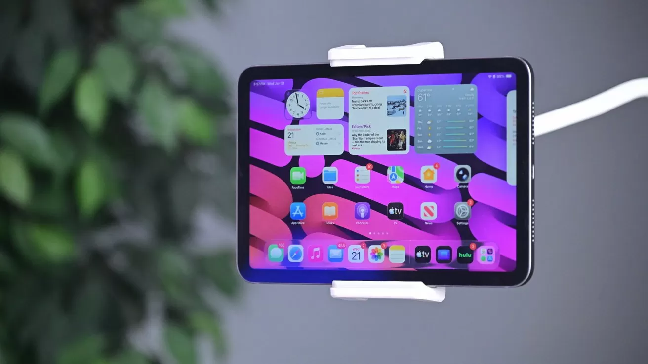 Tablet in a white clamp mount displaying a colorful pink and purple wavy home screen with app icons and widgets, set against a blurred indoor background with green plant leaves
