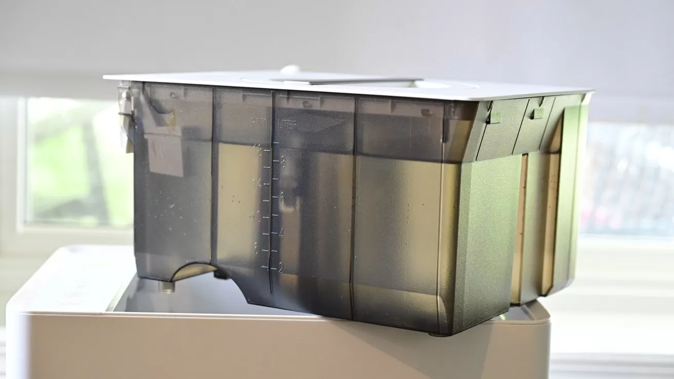 Transparent plastic water tank with visible liquid level markings sits on top of a white appliance, in front of a bright window with soft natural light