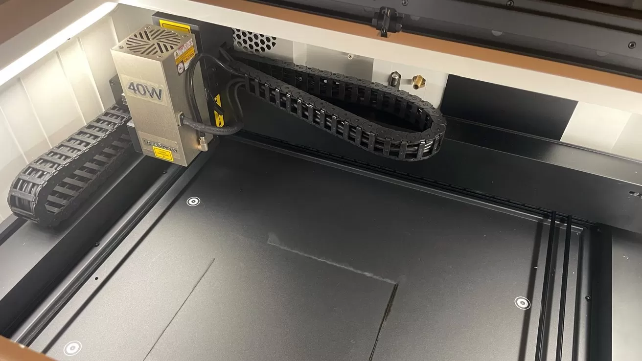 Interior of a laser cutter showing a 40W laser head, cable chain, and large empty black workbed under bright lighting