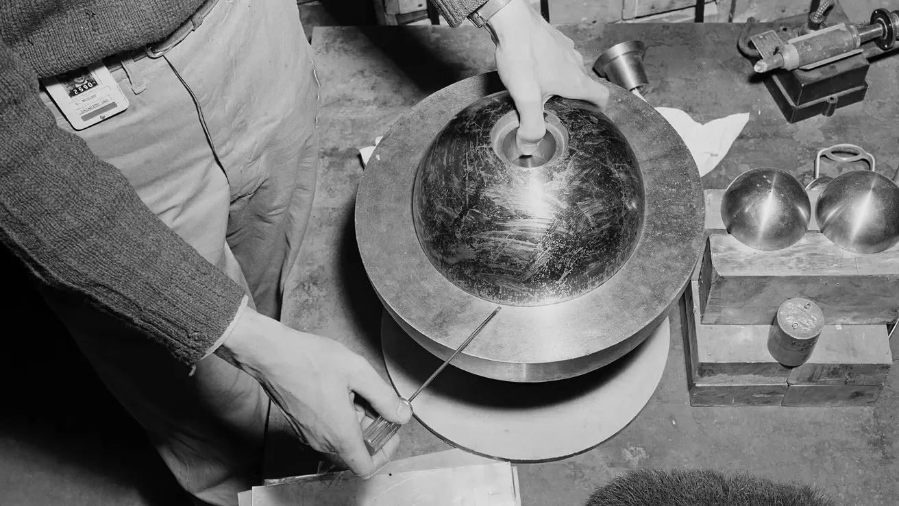 Person assembling a spherical metallic device on a workbench, hands adjusting components around the central core, with additional metal hemispheres and tools arranged nearby in a workshop setting