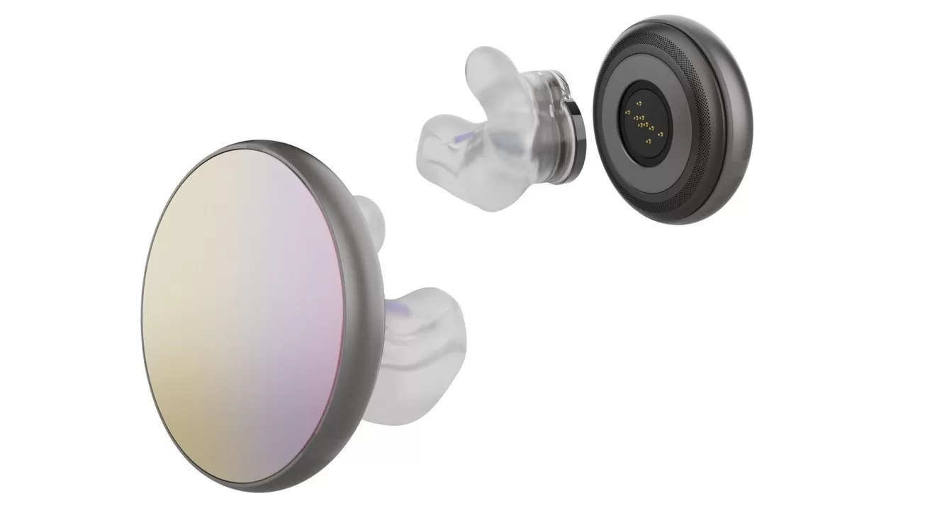 Modern round wireless earbud shown in exploded view, revealing reflective outer face, clear silicone ear tip, and small internal component with gold charging contacts on a white background