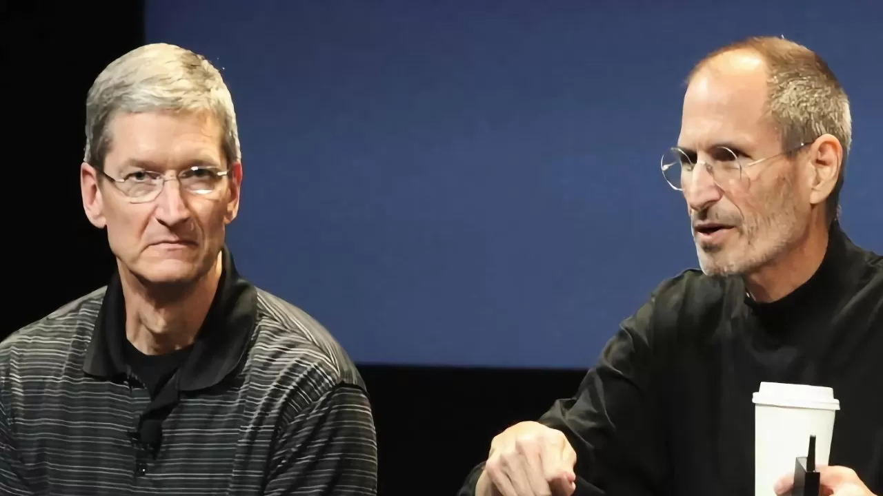 Two middleaged men with short gray hair sit onstage against a dark blue background, one speaking while holding a cup, both wearing glasses and dark casual tops
