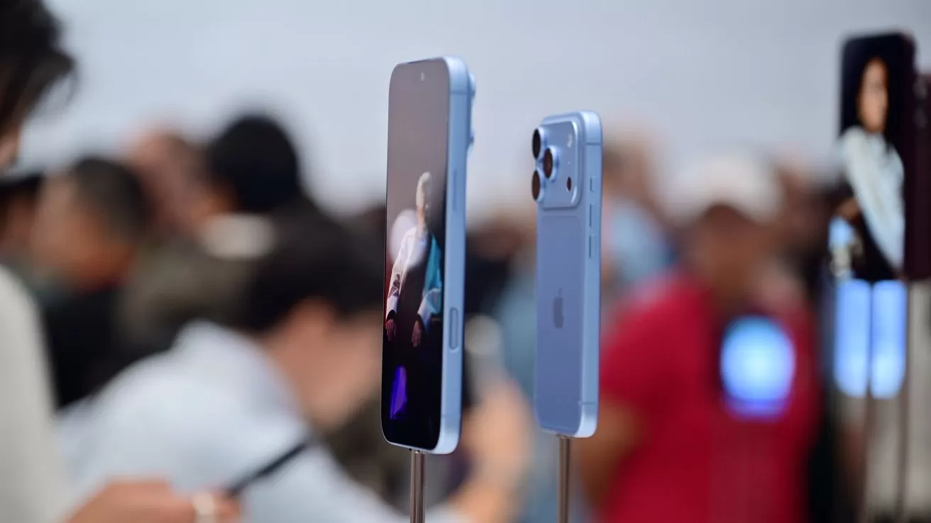 Light blue smartphones on display stands at a busy tech event, screens lit and backs visible, with a blurred crowd of people and lights in the background