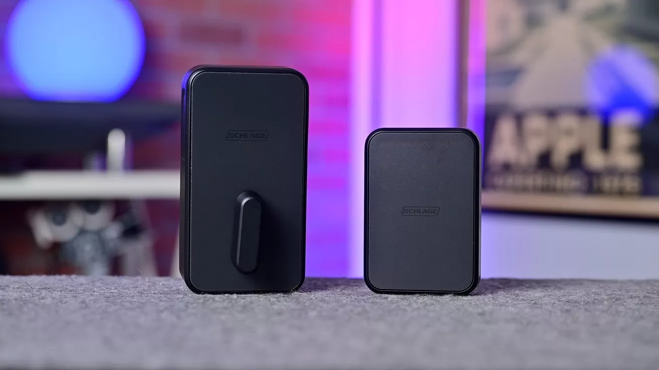 Two black rectangular tech devices on a gray surface, one larger with a protruding clip, against a softly blurred background with colorful lights and a partially visible Apple poster