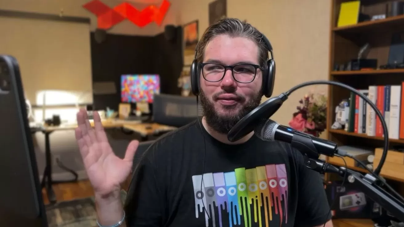 Bearded man with glasses and headphones speaking into a microphone, wearing a colorful T-shirt, sitting in a home office with computer screens, bookshelf, and soft lighting in the background