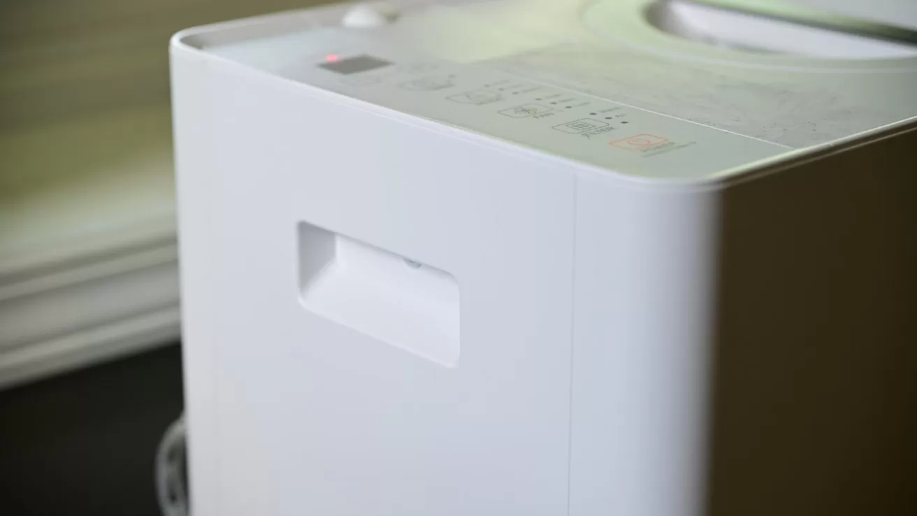Close-up of a modern white appliance, possibly a washing machine or dehumidifier, showing a side handle and top control panel with buttons, near a softly lit window background