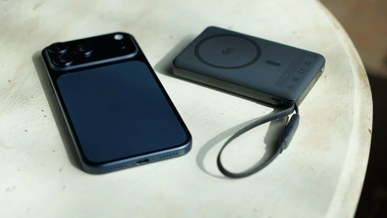 iPhone 17 Pro Max lying face down beside a slim magnetic power bank with wrist strap on a round light-colored table, softly lit by natural light