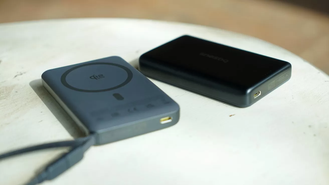 Two slim portable power banks on a round white table, one gray with wireless charging symbol and connected cable, the other black with a visible USB port, softly lit.