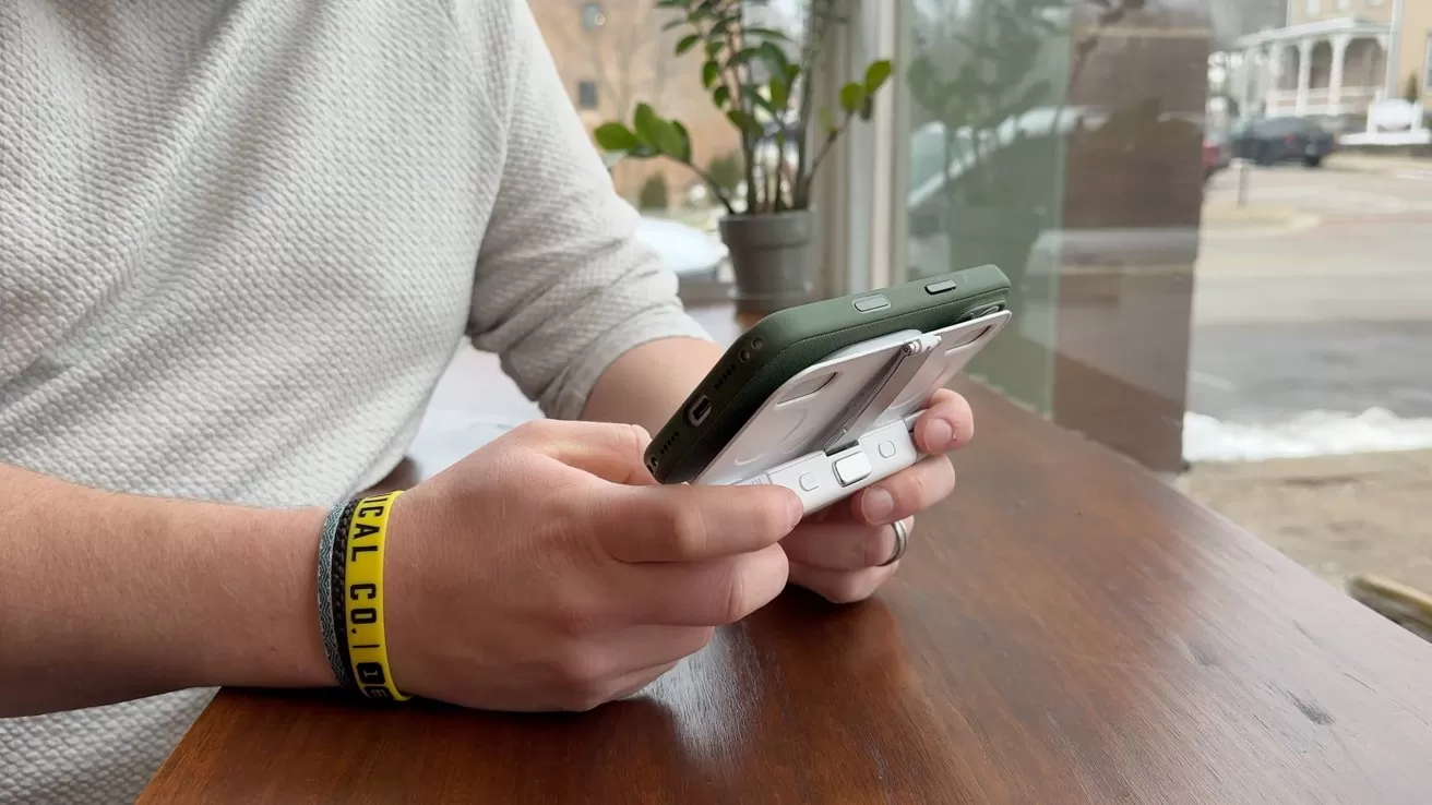 Person wearing a textured sweater holds a green phone with a white gaming attachment, seated beside a window with a potted plant visible.