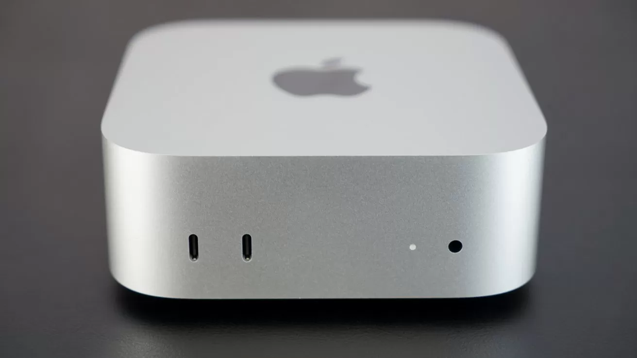 Silver Apple Mac mini desktop computer on a dark surface, showing front ports and small indicator light, with the Apple logo centered on the top