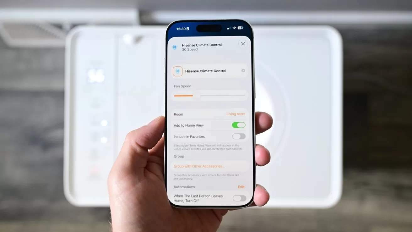 Hand holding a smartphone displaying a home climate control app screen with temperature settings and toggles, in front of a blurred white household device on a wooden floor