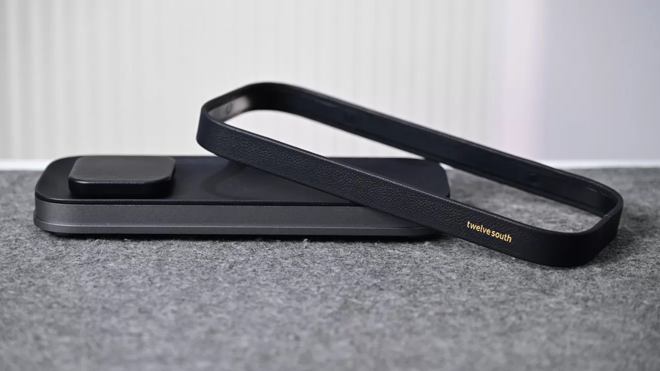 Slim black wireless charging pad on a gray fabric surface, with a matching black rectangular frame resting diagonally on top, featuring small gold text on one side