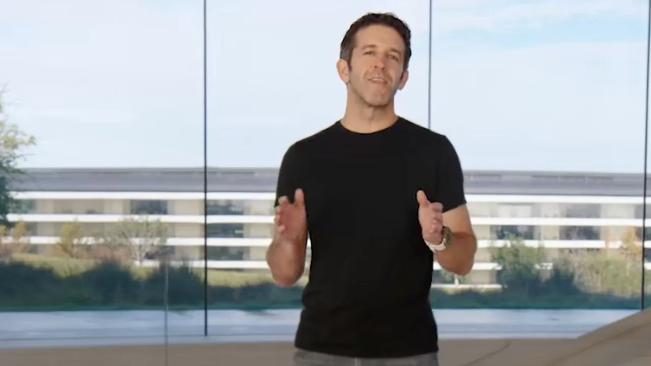 Man in a black T-shirt speaks while gesturing, standing indoors before tall glass windows overlooking a modern circular office building and landscaped greenery under a bright sky