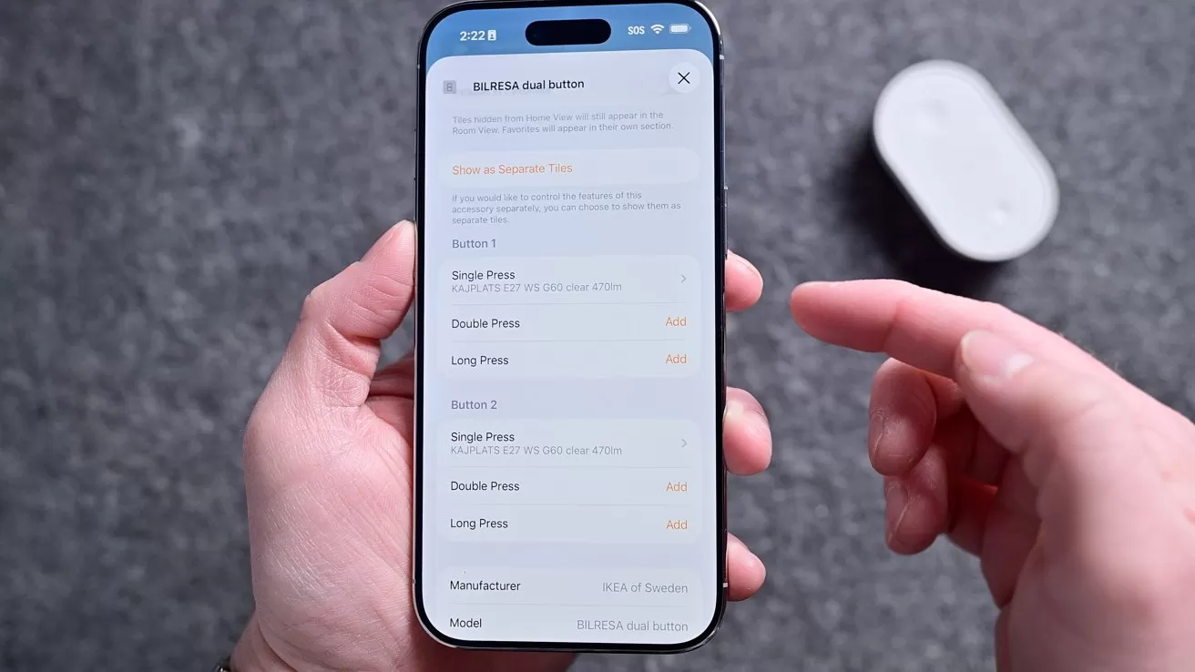 Person holding a smartphone showing a settings screen for customizing button presses, while their other hand points at the display; a small white gadget rests blurred in the background