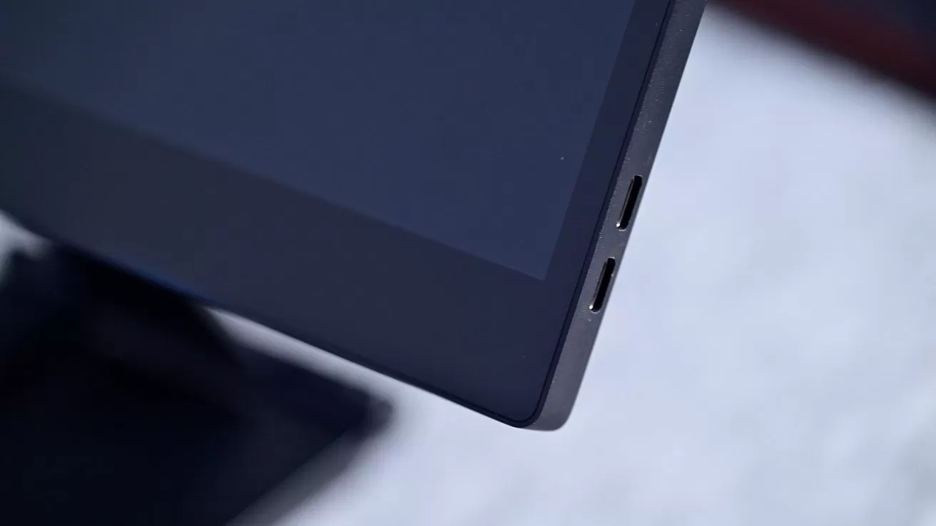 Close-up of a black computer monitor corner featuring two USB-C ports against a blurred gray background.