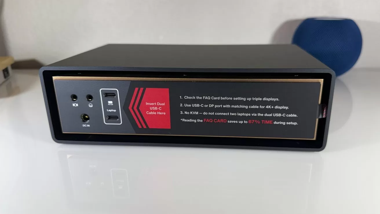 Black rectangular docking station front panel showing audio jacks, DC input, two USB-C laptop ports, and a red label with setup instructions, resting on a white desk with blurred background.